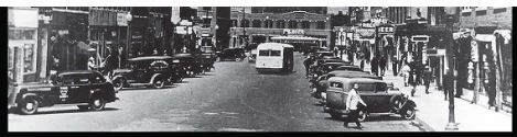 Panoramic view of a thriving Black commercial district street, showing storefronts, automobiles, and pedestrians